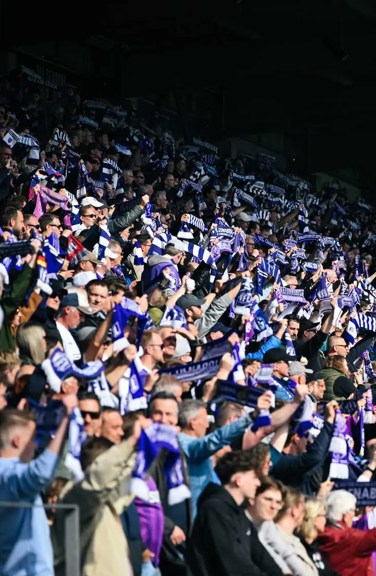 ING_7132 (1)_hoch Volle Tribüne: Fußballfans mit Schals im Stadion.